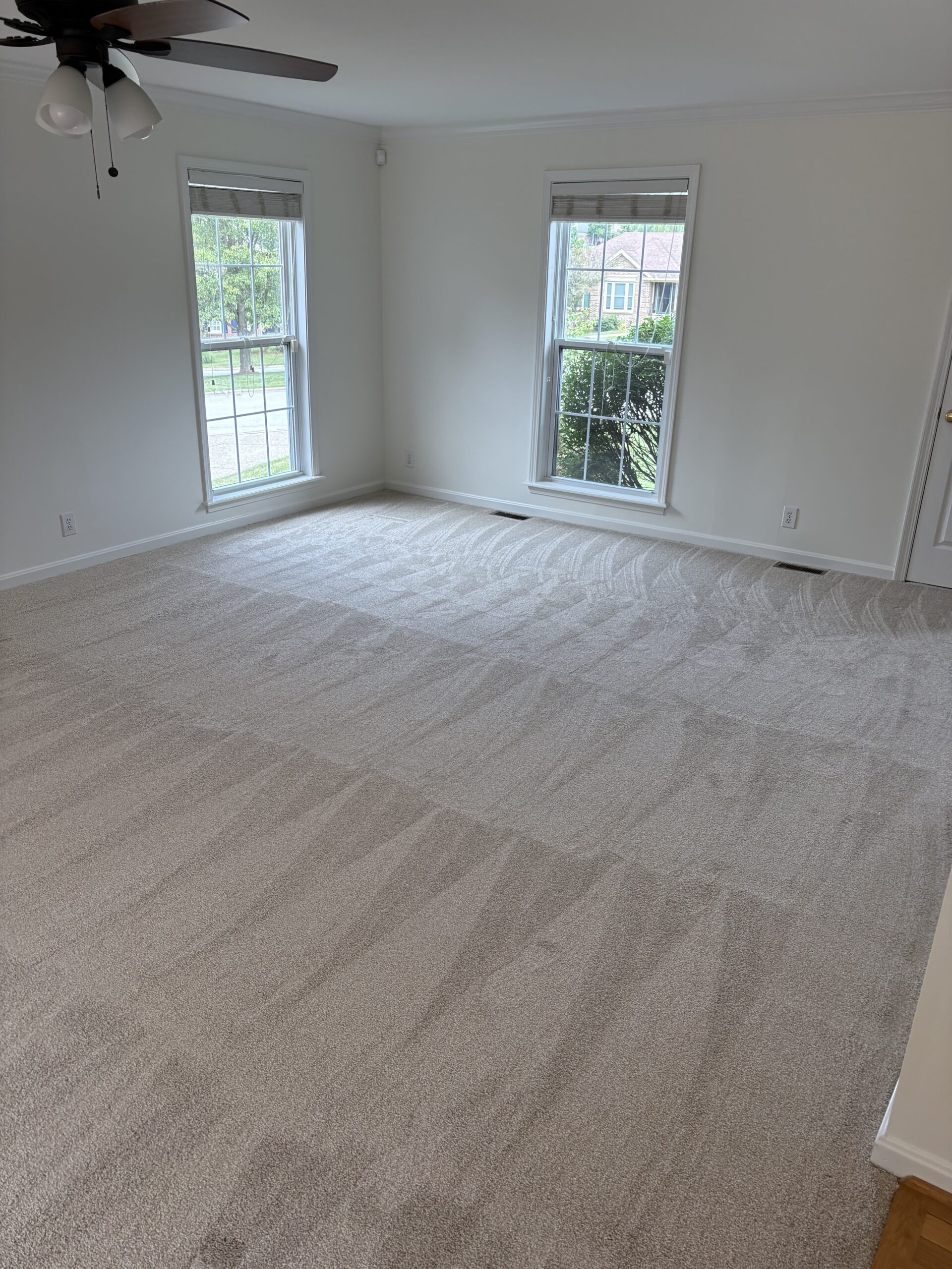 Empty room with clean, light-colored carpet, two large windows letting in natural light, white walls, and a ceiling fan.