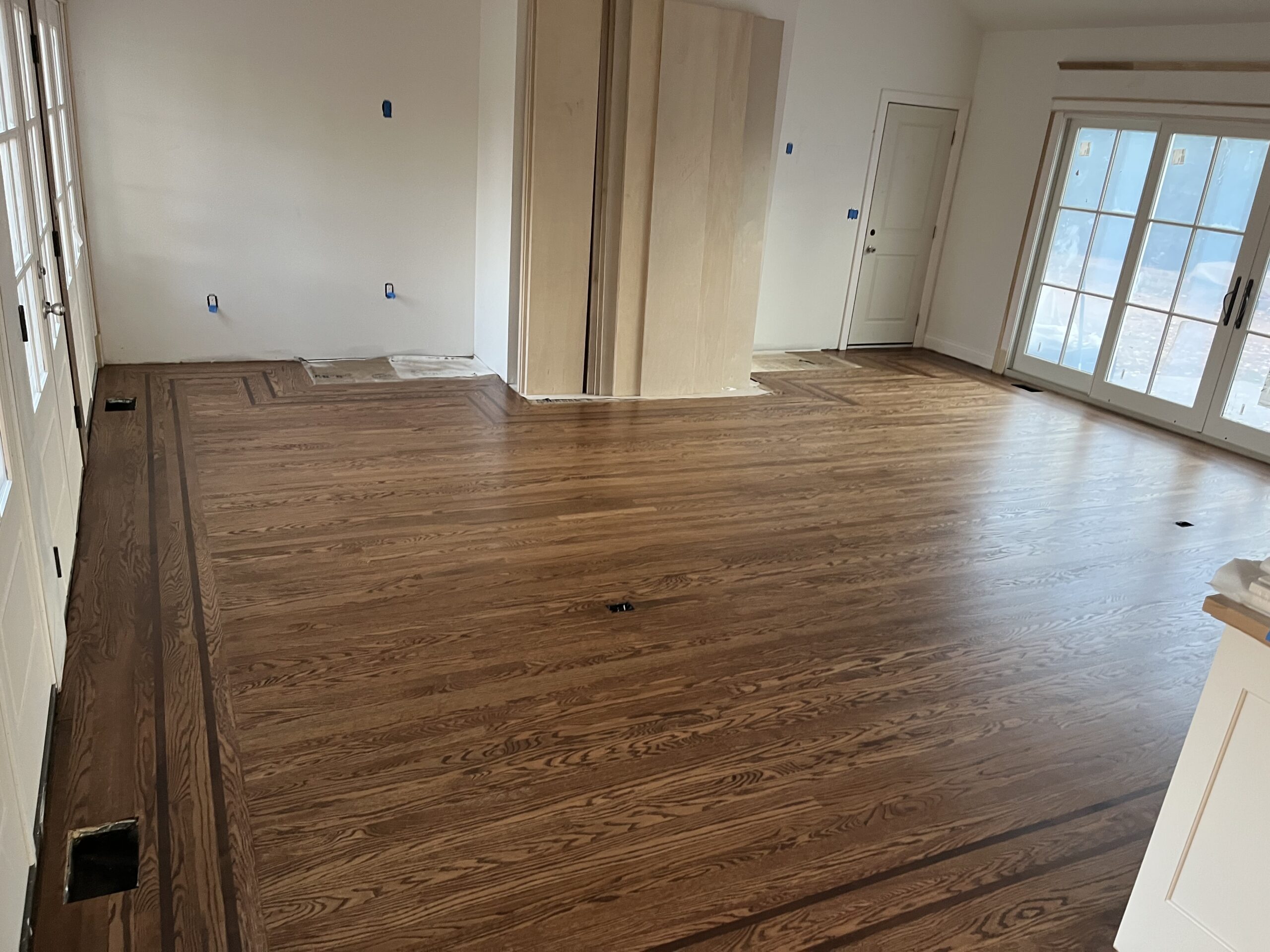 A room with newly installed hardwood flooring featuring a decorative border, unfinished walls, and covered windows and outlets prepared for painting after expert hardwood floor sanding.