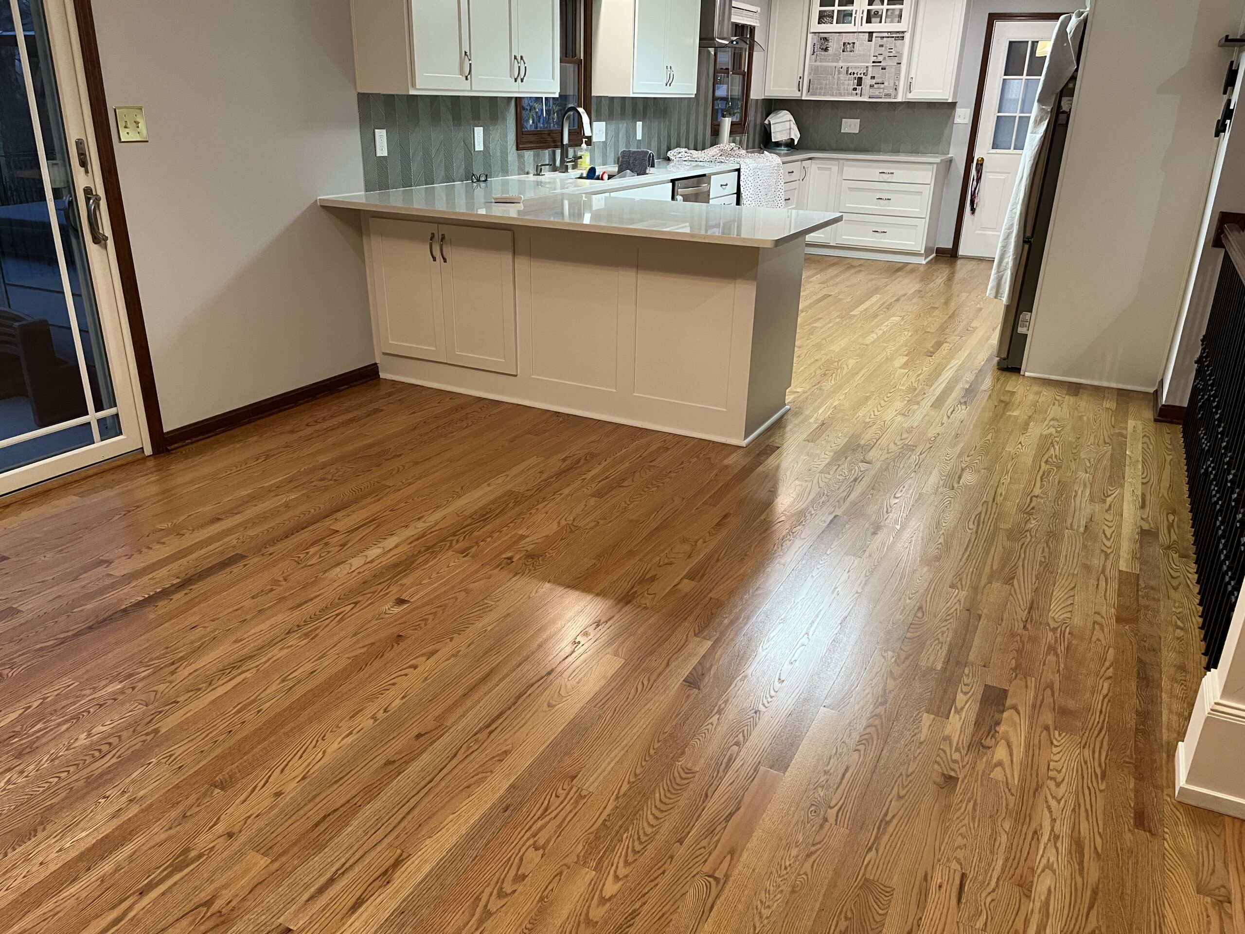 A kitchen with white cabinets and an island is adjacent to a spacious area with polished wooden floors and sliding glass doors.