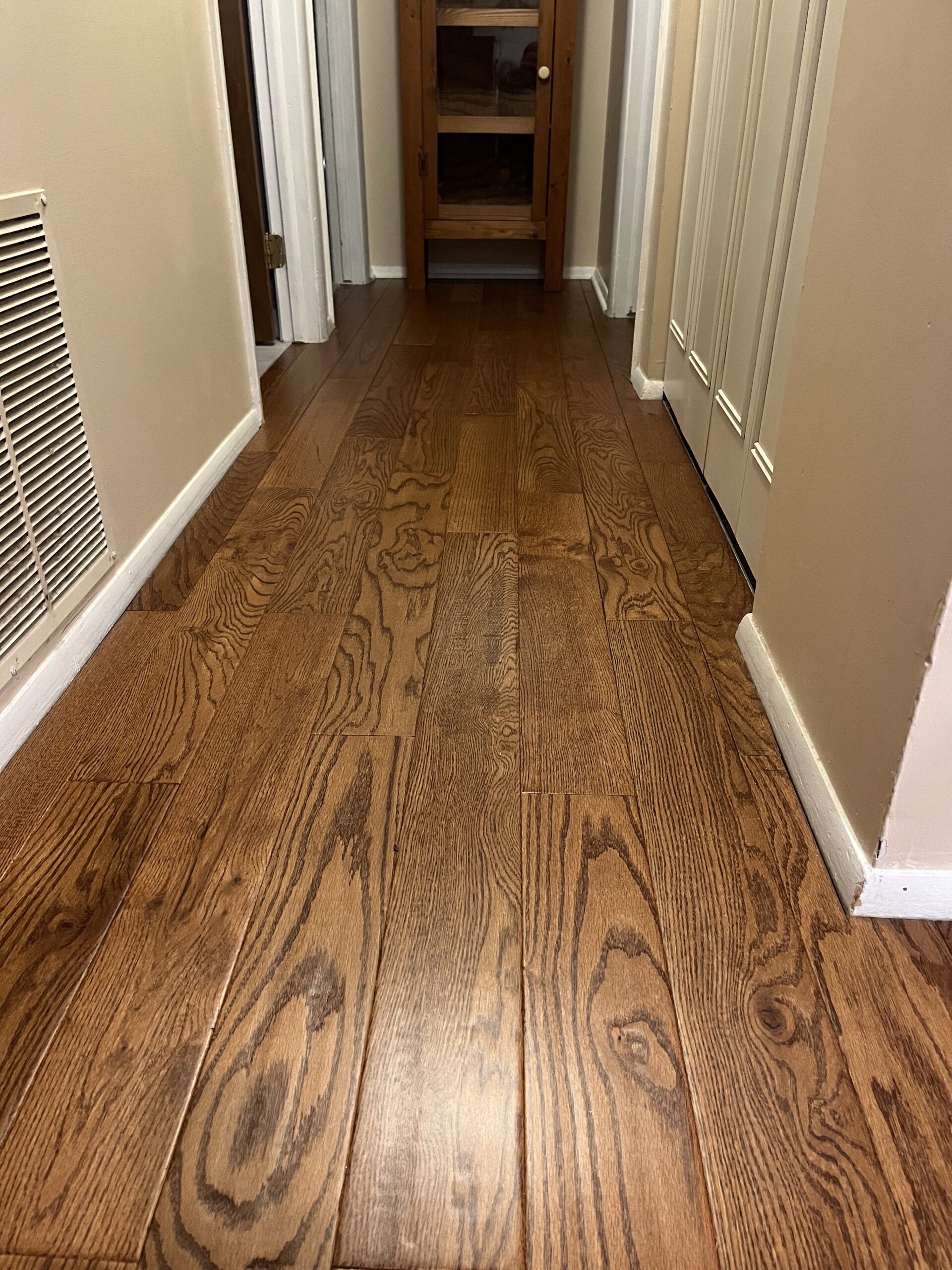 A hallway with light brown wood flooring, cream-colored walls, and white baseboards. Doors are along the sides and a wooden cabinet is at the end of the hall.
