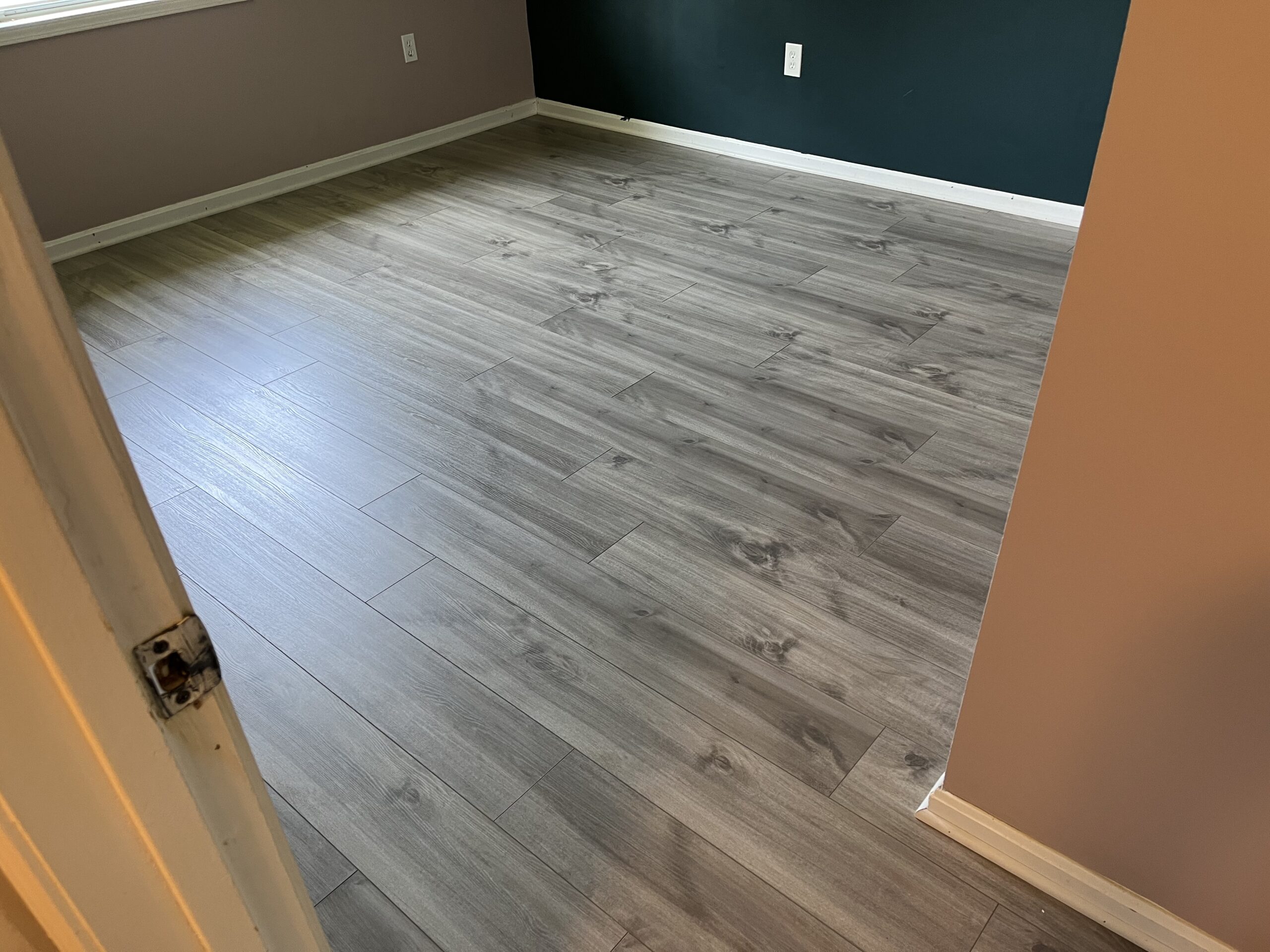 A room with light gray wood-look laminate flooring, beige and dark green painted walls, white baseboards, and an electrical outlet visible on the far wall.