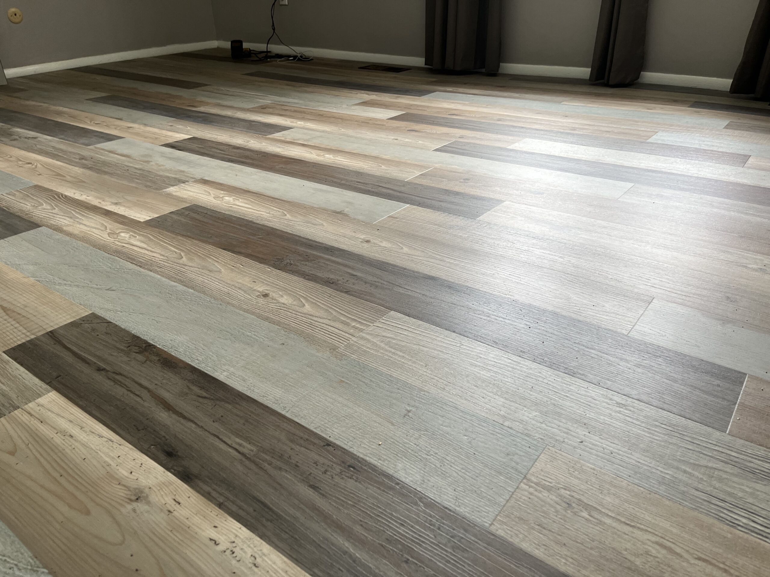 A room with light and dark wood-patterned laminate flooring, bare walls, and windows with dark curtains. An electrical cord is visible against the far wall.