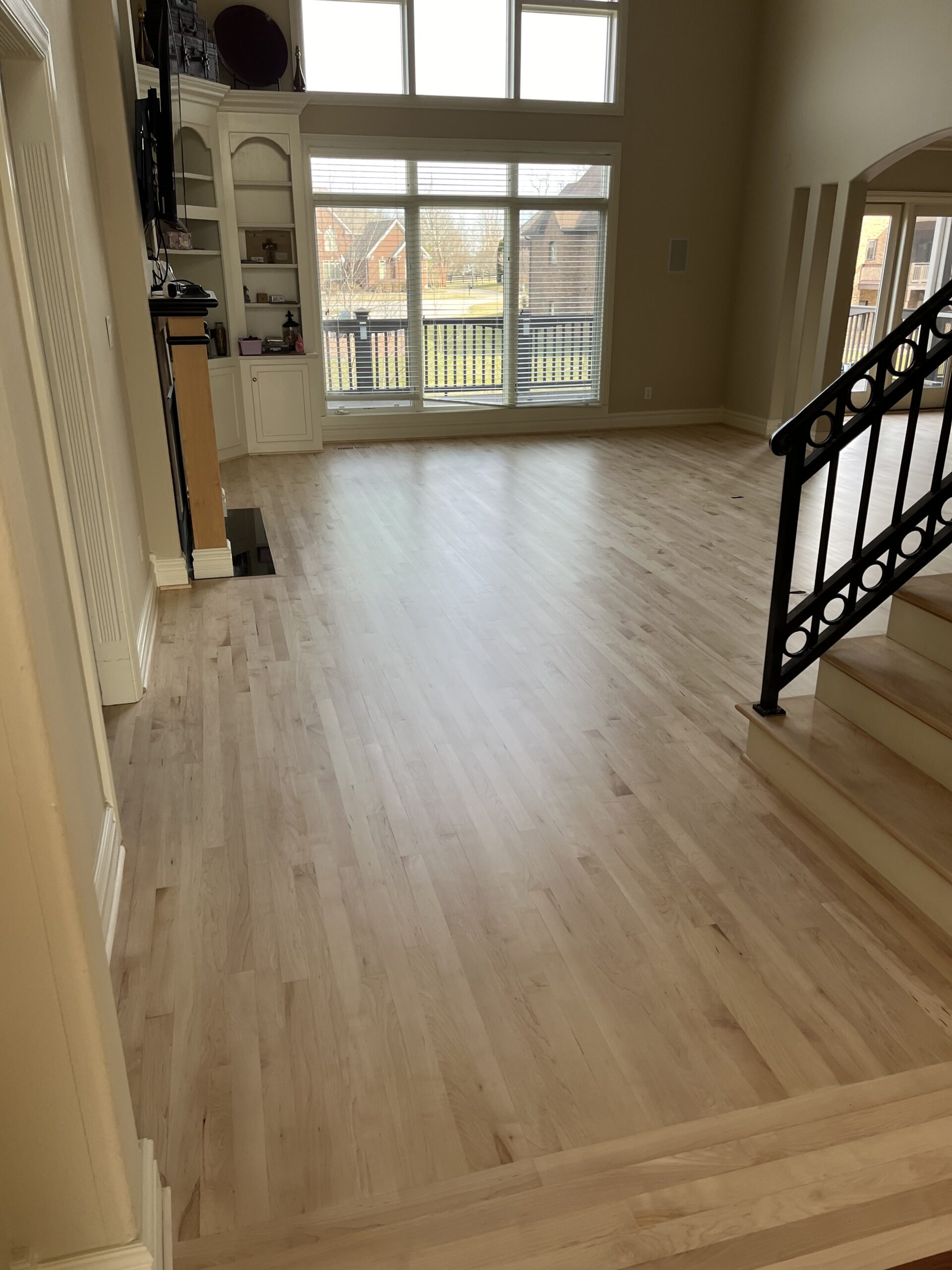 Spacious living room with light wood flooring, large windows overlooking a backyard, built-in shelves, and a staircase with a black railing.